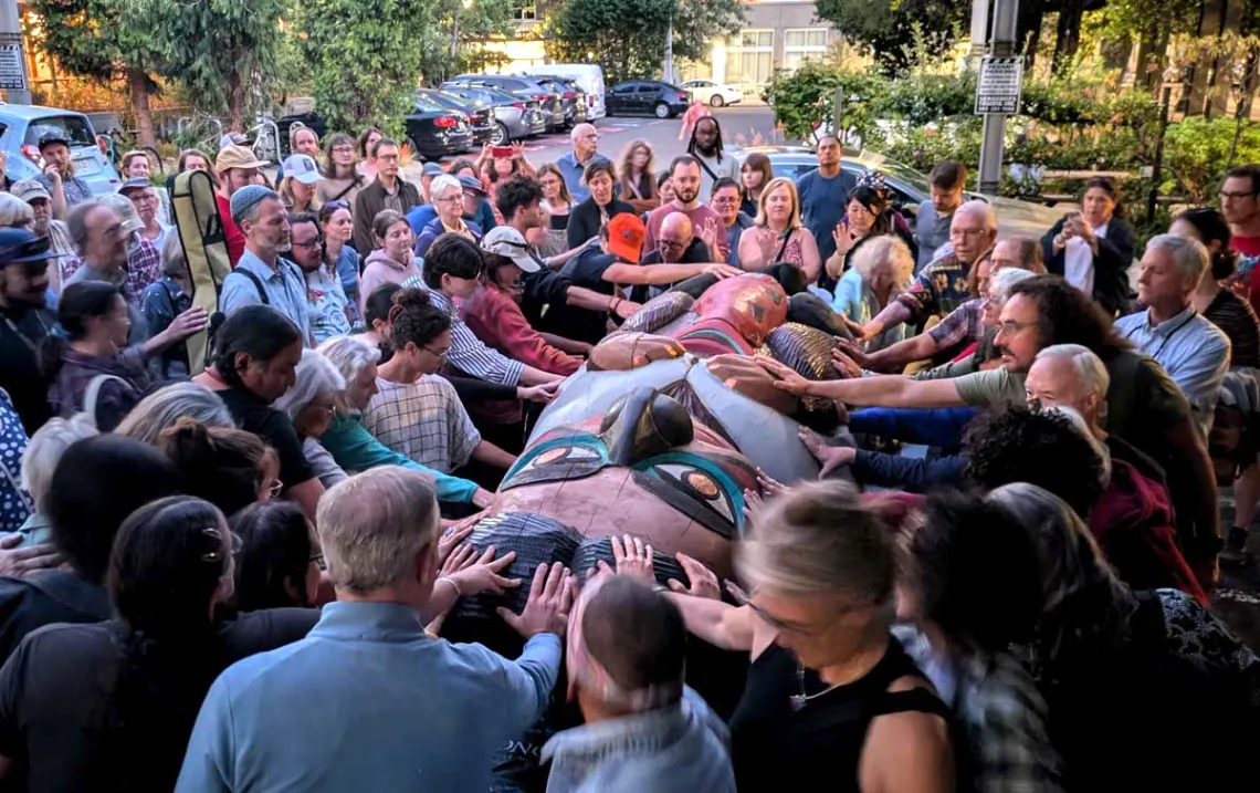 A group of people crowded around a large carved totem laying on a truck trailer.