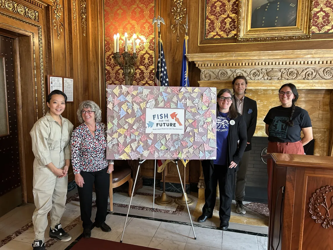 Press conference speakers stand in front of a display of origami fish.