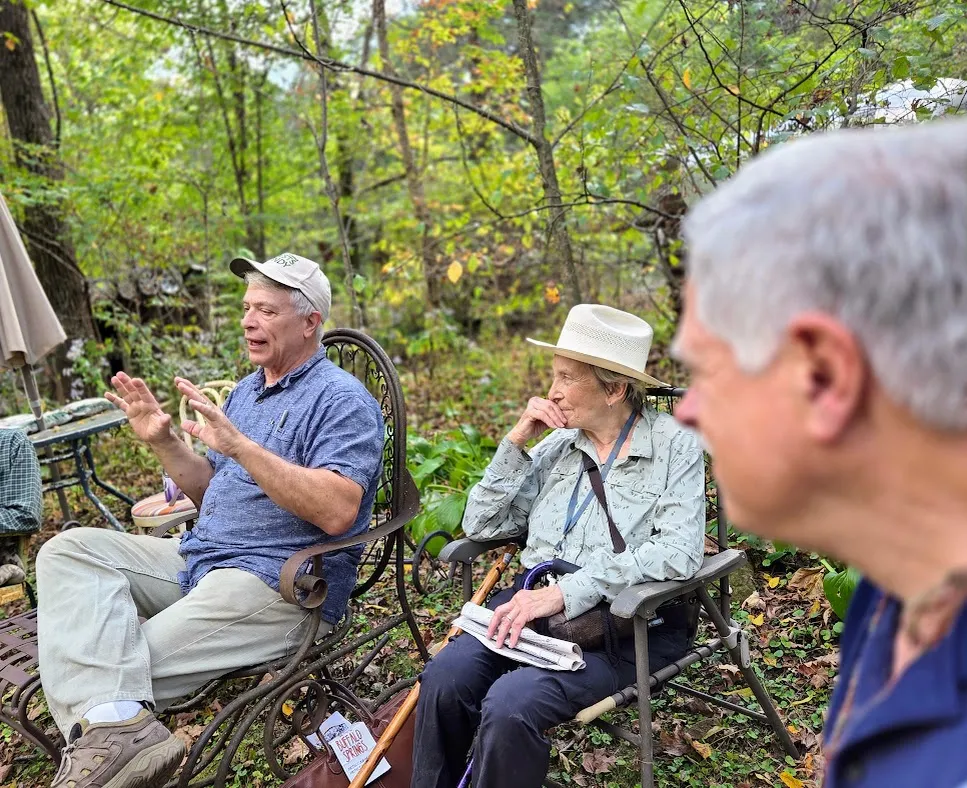 Three people sitting on camp chairs in a woods. One of them is talking and gesturing with their hands. The other two people are listening to him.