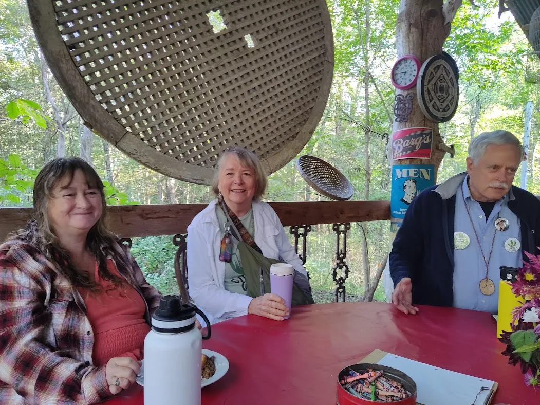Three people sat at a red table outdoors on a wooden deck or porch. There are trees behind them. They are smiling and in conversation with each other.