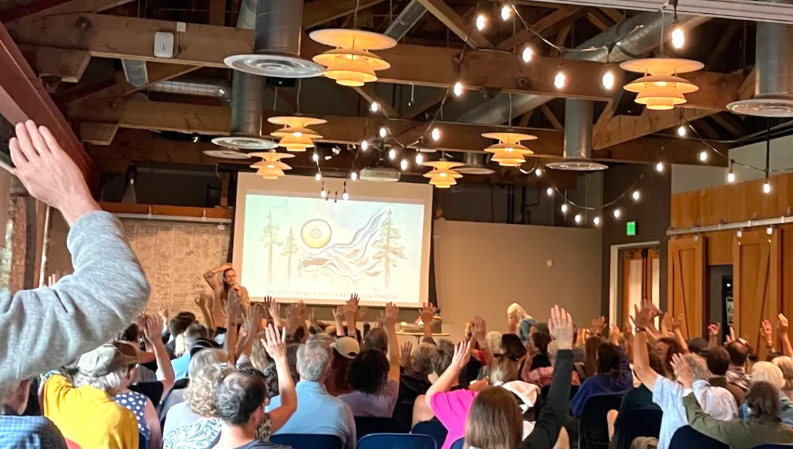 Photo of a medium sized room filled with seated people facing a presenter at the front of the room. The presenter and many in the audience have their hands raised.