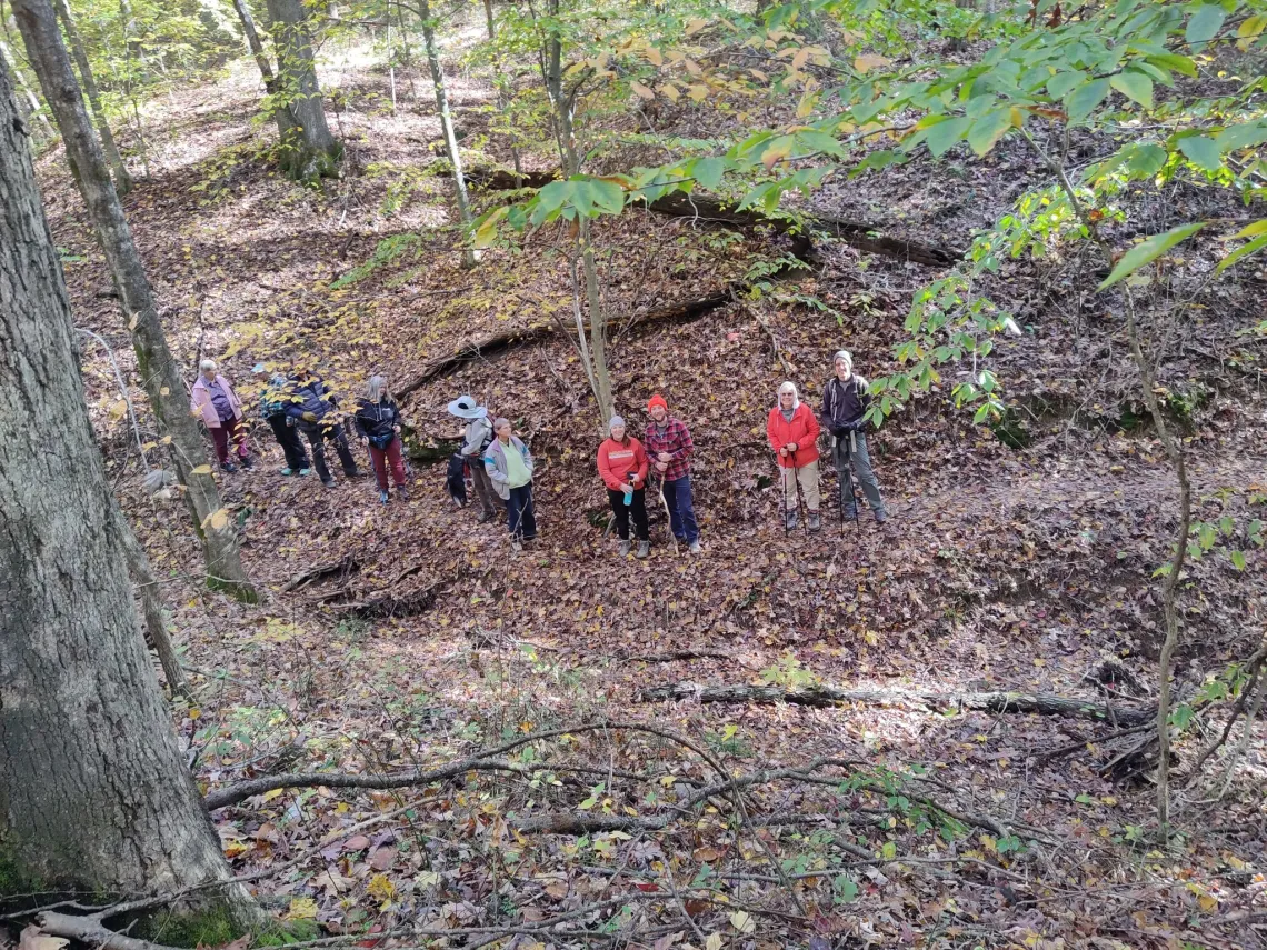 Looking down at a group of around 10 folks in a forest. They are dressed for cool-ish weather. They are looking up for the photo. The ground is covered in brown leaves but some of the trees around still have some green leaves on them 