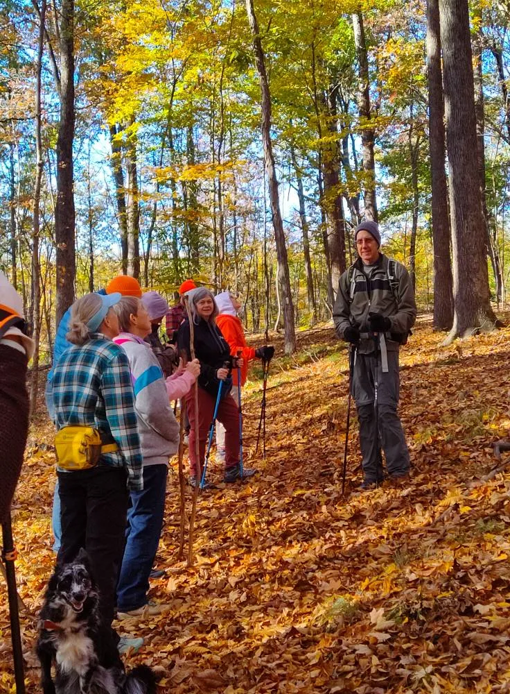 A group of around 7 people in a forest. One person is standing slightly apart from the main group and is speaking to them. The ground is covered in brown and orange leaves. The leaves remaining on the trees are green and orange. The sky is blue.