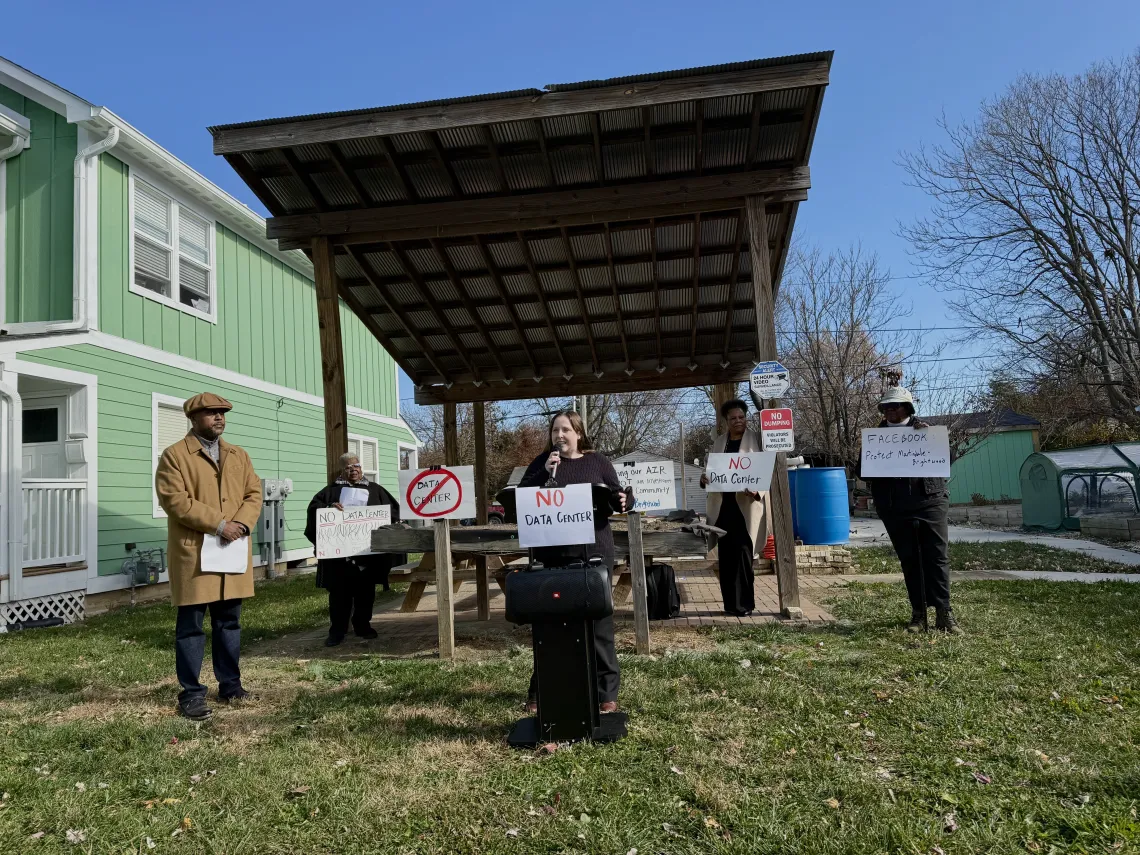 Five people stood outdoors in a small neighborhood park. One person is standing forward of the others and is speaking through a microphone. There is a sign on the microphone stand saying NO data center. The other people in the photo are all holding signs too. There area  few trees in the background which have shed their leaves. The sky is blue.