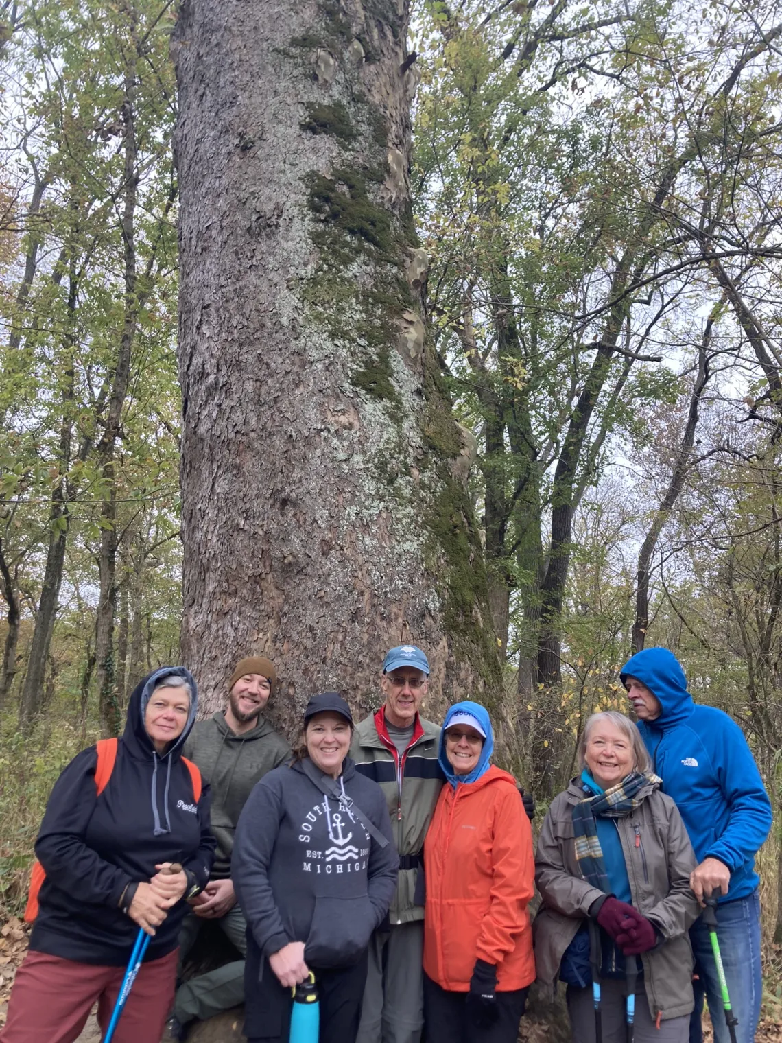 A group of 7 people around the base of a tall tree in the woods. They're dressed for cool weather. Some are wearing caps and have walking poles. They are all smiling.