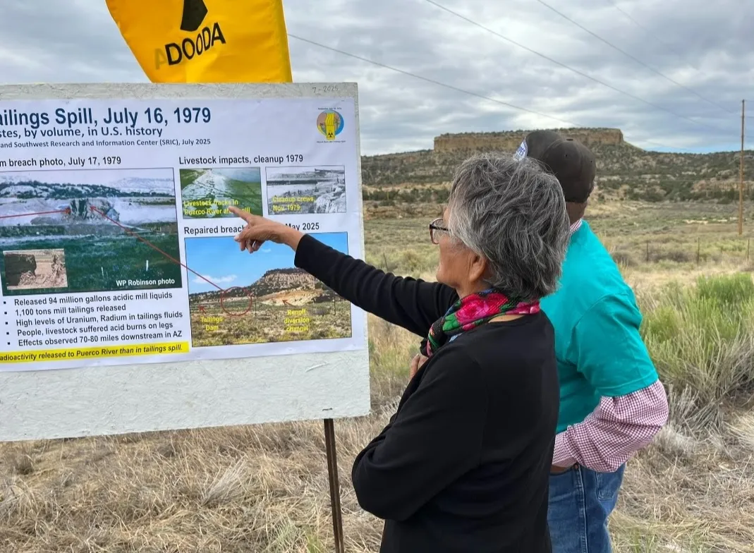 Photograph from the Church Rock commemoration event.  Image shows an individual with their back turned to the camera pointing at a poster which displays information on the 1979 Uranium Spill.