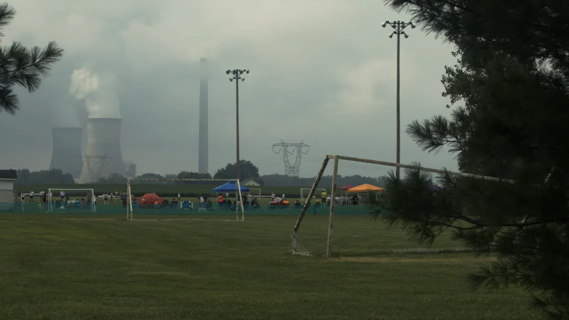 Coal plant towers billowing out smoke in background. In the foreground is a sports field with soccer goals with some players gathered at the far end. The sky is a smokey gray.