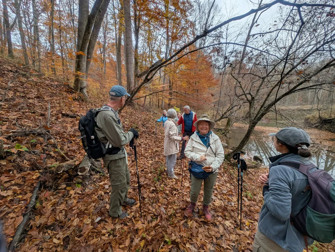 A group of around 6 people in the woods. They are dressed for cooler weather. There is a small body of water nearby and brown and orange leaves on the some of the trees and on the ground.