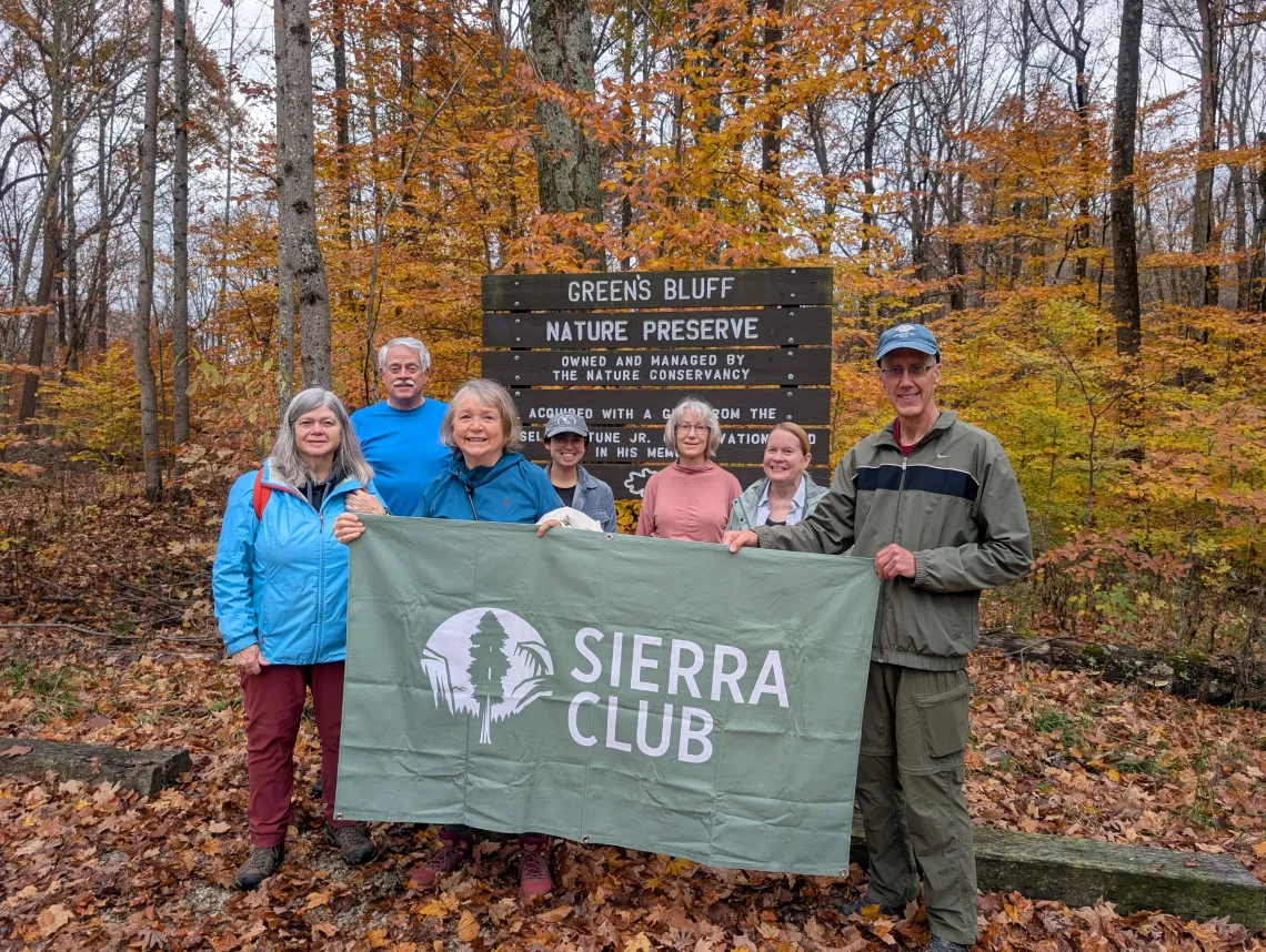 A group of 7 people at a trail head. They are holding a green Sierra Club banner and dressed for the cooler weather. The leaves on the ground and on the trees are brown and yellow.