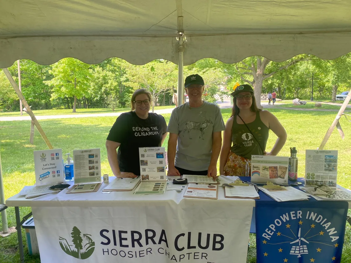 Three people stood behind a table at an outdoor event. They are all smiling. The table has banners on it - one says Sierra Club Hoosier Chapter and the other says Repower Indiana.