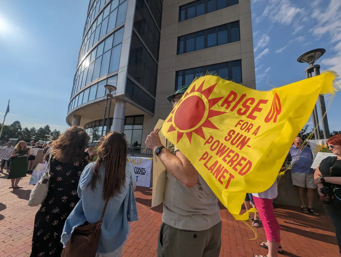 People at a rally on a sunny day outside a glass-fronted building. There are three people in the foreground with their backs to the camera. One is holding a large yellow flag with a red sun and writing also in red saying RISE FOR A SUN-POWERED PLANET