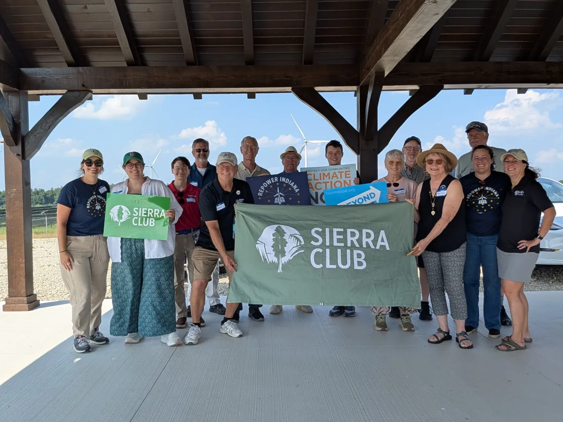A group of fourteen people standing outside. Some of them are holding signs and banners saying "Sierra Club" and "Repower Indiana". The sky behind them is blue with small fluffy clouds, and a couple of wind turbines are also in the background.