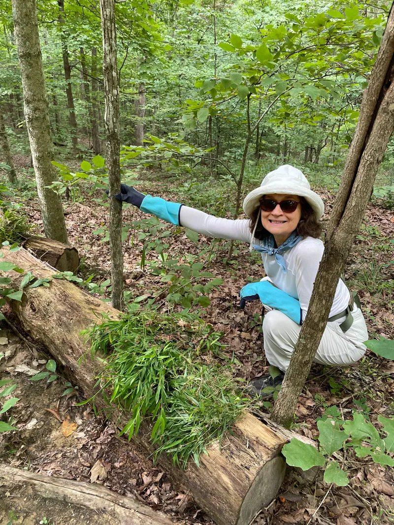 A person kneeling down in the woods holding onto a sapling with one hand. They're wearing gloves, a white cap and dark sunglasses. They are smiling.