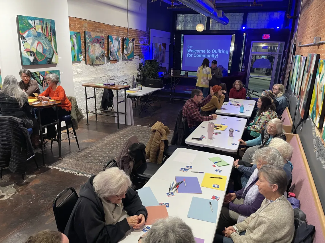 People in small groups of 5 sit at tables with paper and markers at the Quilting for Community Kick-off.