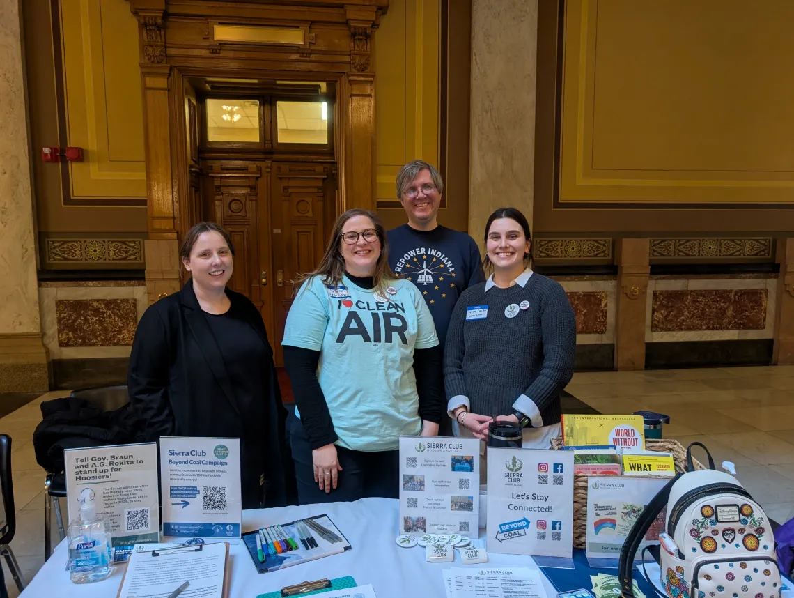 Four people tabling indoors. There is lots of information on the table including flyers and petitions. One of the people is wearing a shirt saying I heart clean air. Another is wearing a shirt saying Repower Indiana. All four people are smiling.