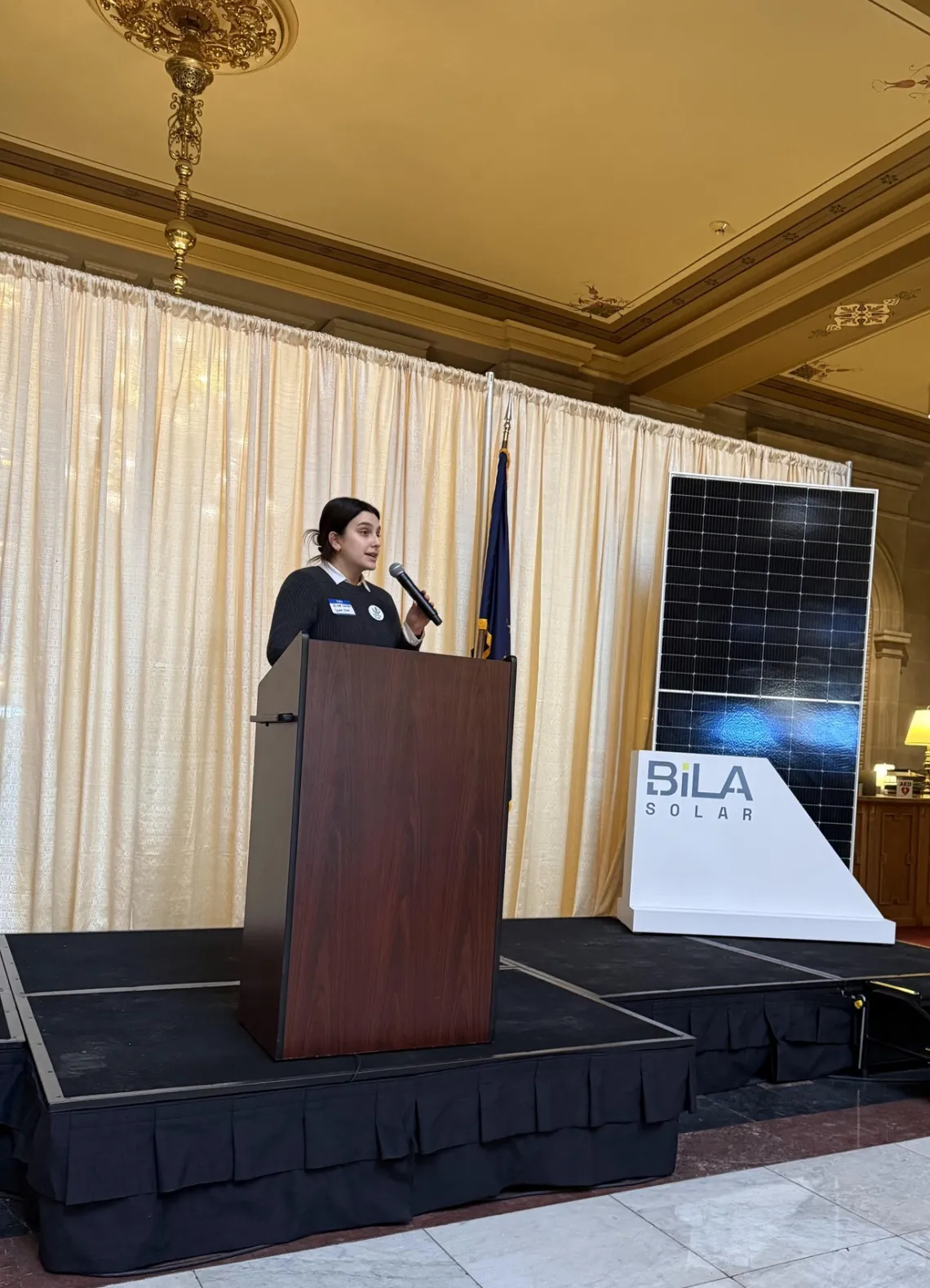 A person with long brown hair pulled back standing behind a podium at a small indoor stage, speaking into a microphone