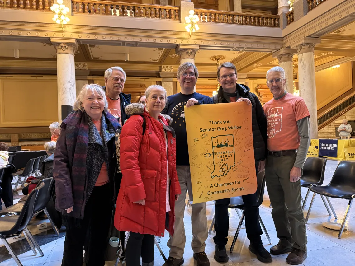 Six people standing indoors at the Statehouse. There are pillars behind them and chairs spread out in the room for attendees. Two of the people are holding up a yellow poster with an outline of the Indiana map on it. The poster says Thank you Senator Greg Walker. A champion for Community Energy. The people in the photo are smiling.