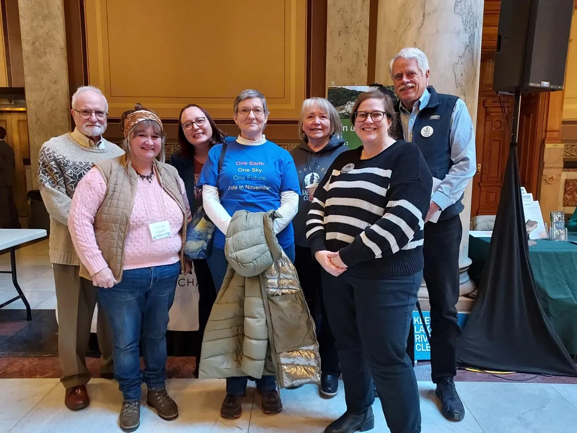 Seven people of varying ages standing indoors at the Statehouse. They are all smiling for the photo.