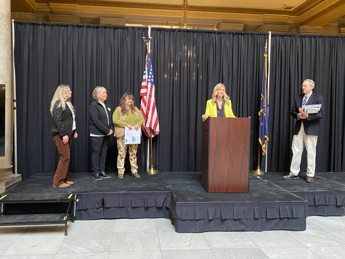 Five people on a low stage indoors with black curtains behind them and an American flag. One of them is speaking behind a podium and wearing a yellow jacket. The other three people are listening.