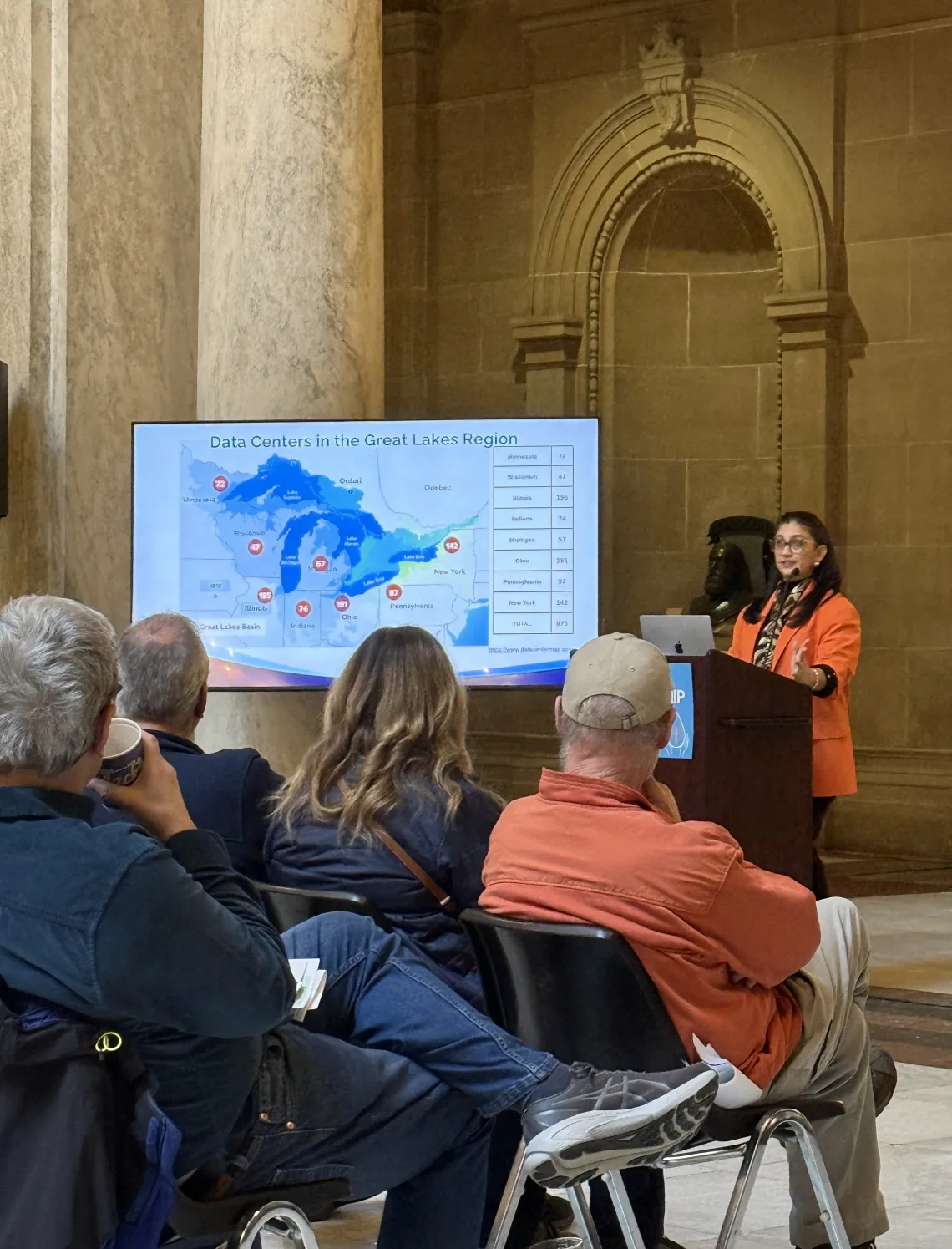 A person with dark hair and an orange jacket speaking at a podium indoors. There is a presentation showing a map of data centers in the Great Lakes region. There are people seated in chairs with their backs to the camera watching the person talk.