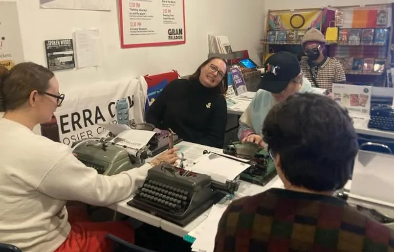 Five people indoors typing on old fashioned typewriters. Some are wearing respirator masks. There are books for sale in the background.