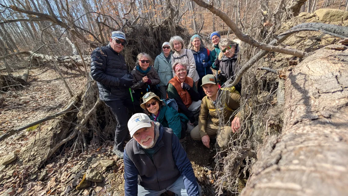 A group of around 11 people outdoors in a forest. They are all standing or kneeling between the bases of two large fallen trees. They are all dressed for cooler weather. Some are wearing hats and glasses. They are all smiling.