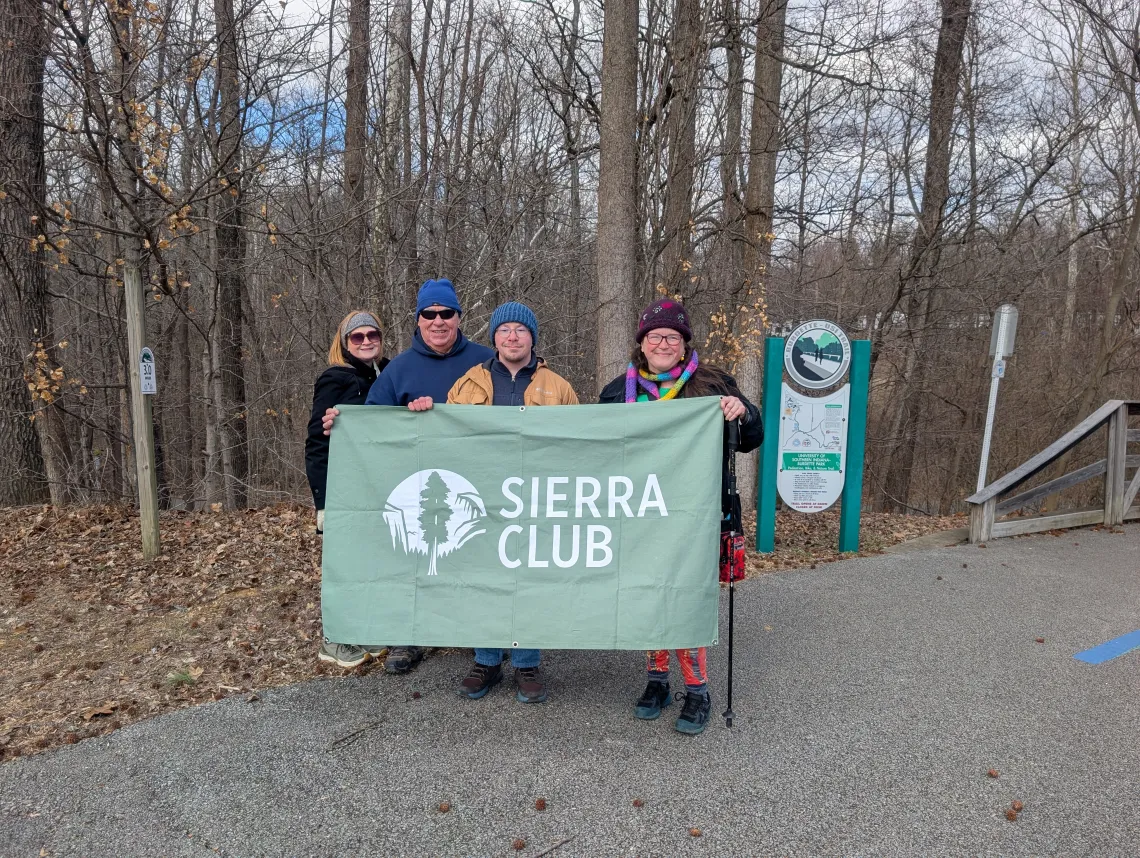 Four people at a trail head holding a green Sierra Club banner. They are dressed for the cold weather in coats and hats. The trees behind them are bare and there are brown leaves on the ground.