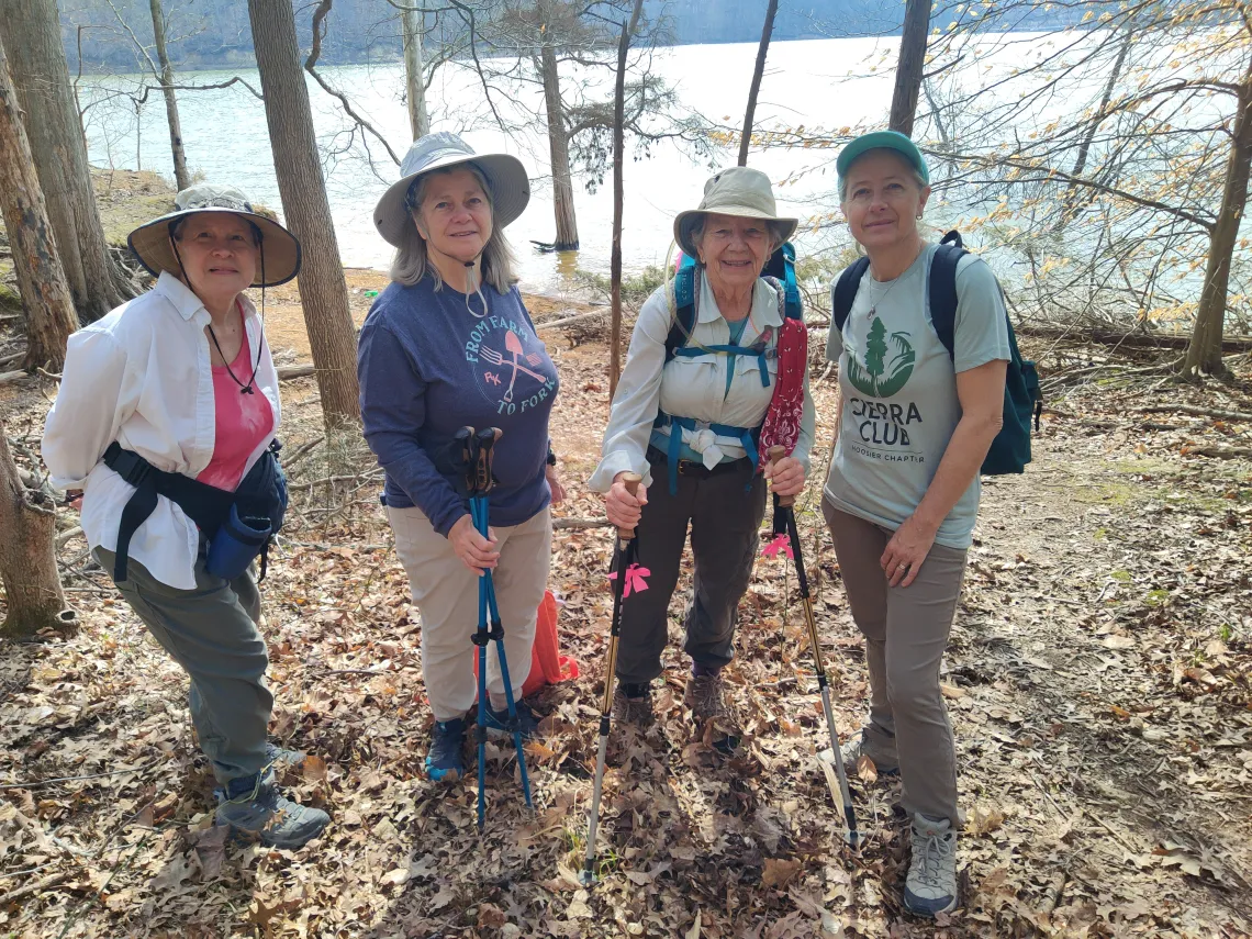 Four people on a hike with a body of water behind them and sparse trees with brown leaves on the ground. They are wearing sun hats and some of them have hiking poles. One of them is wearing a Sierra Club tee.