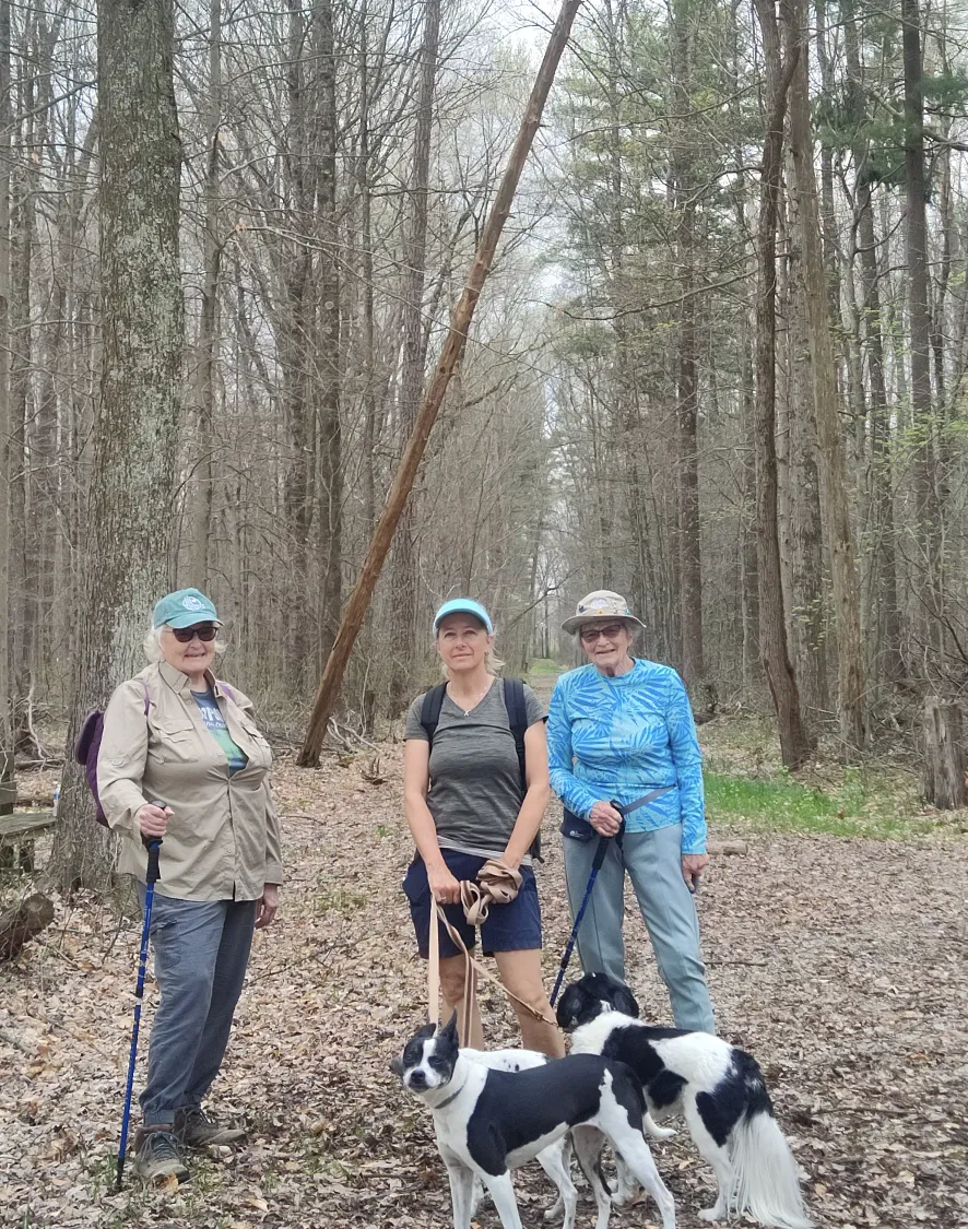 Three people on a leafy trail with tall trees behind them, with two black and white dogs. They are all smiling. 