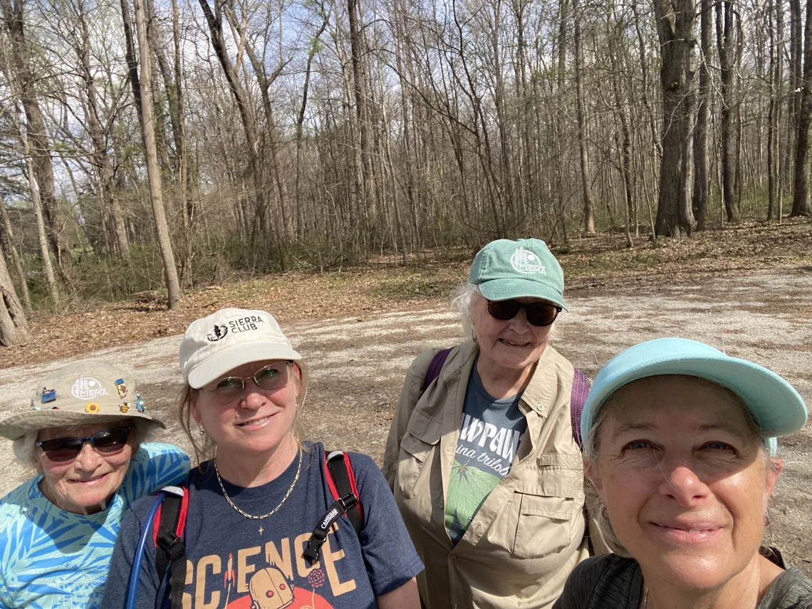 Four people on a hike with trees behind them. They are all wearing hats. Two of the people are wearing sunglasses and one is wearing a t-shirt saying Science and has a Sierra Club logo on their hat. They are all smiling.