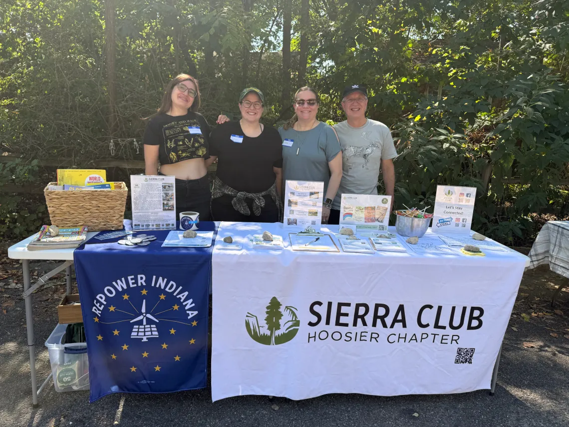 Four people tabling outdoors on a street. There are trees behind them. They are smiling for the photo. On the table there is a white tablecloth with the Sierra Club Hoosier Chapter logo of black text and a green tree graphic, and a smaller blue tablecloth which says Repower Indiana with a white and yellow logo of a wind turbine and solar panels surrounded by a circle of stars. On the table there are books, petitions, stickers, and other info about the Sierra Club.