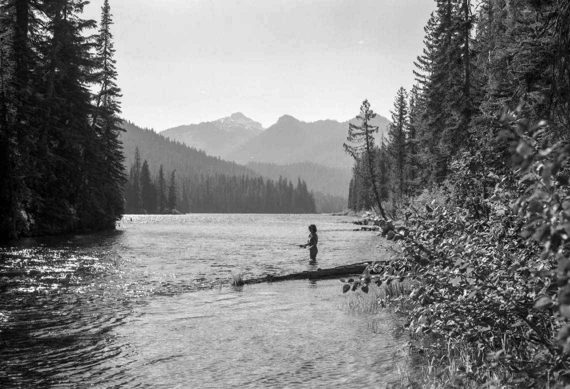 a black and white photo of a woman fishing in a lake surrounded by evergreen trees and mountains