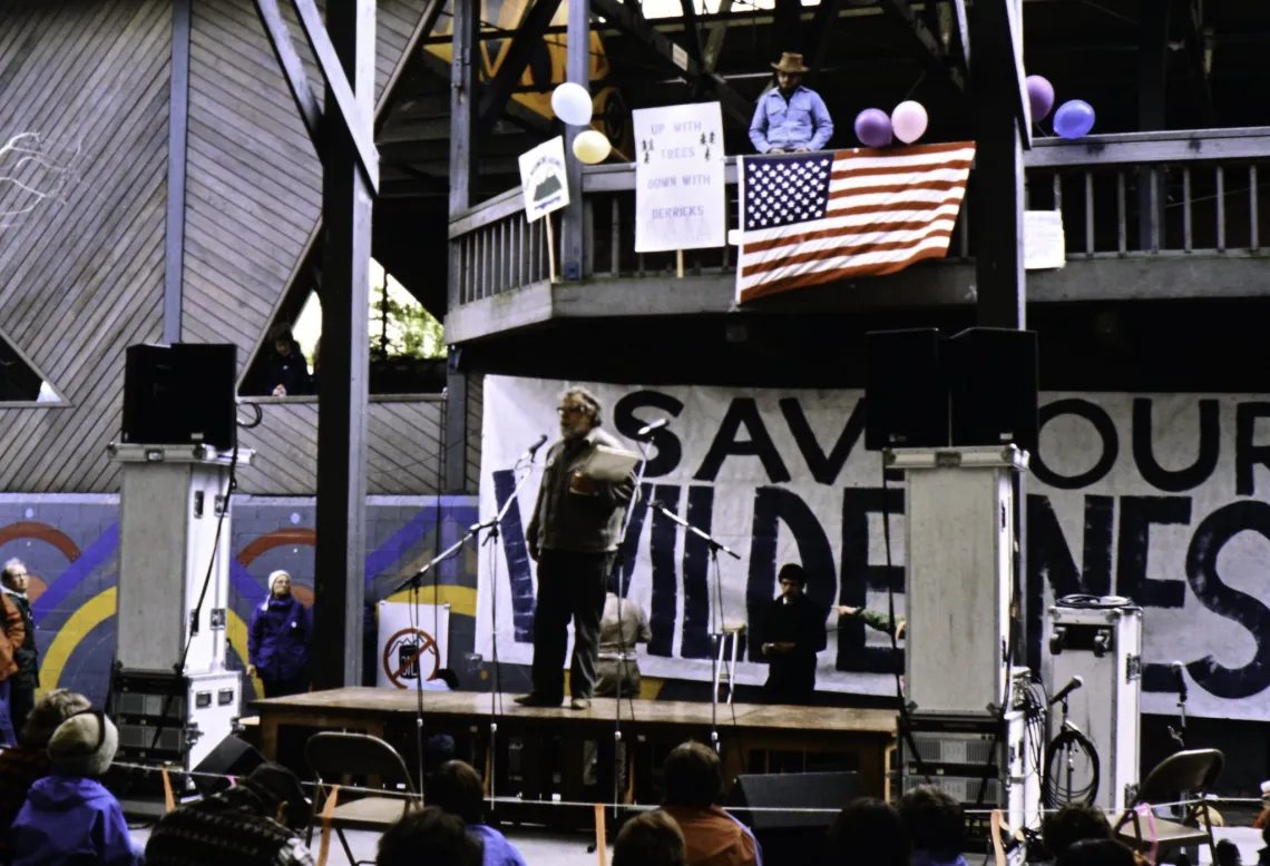 a man, Harvey Manning, stands on a stage in front of a crowd with a large black and white hand painted banner behind him that reads "save our wilderness"