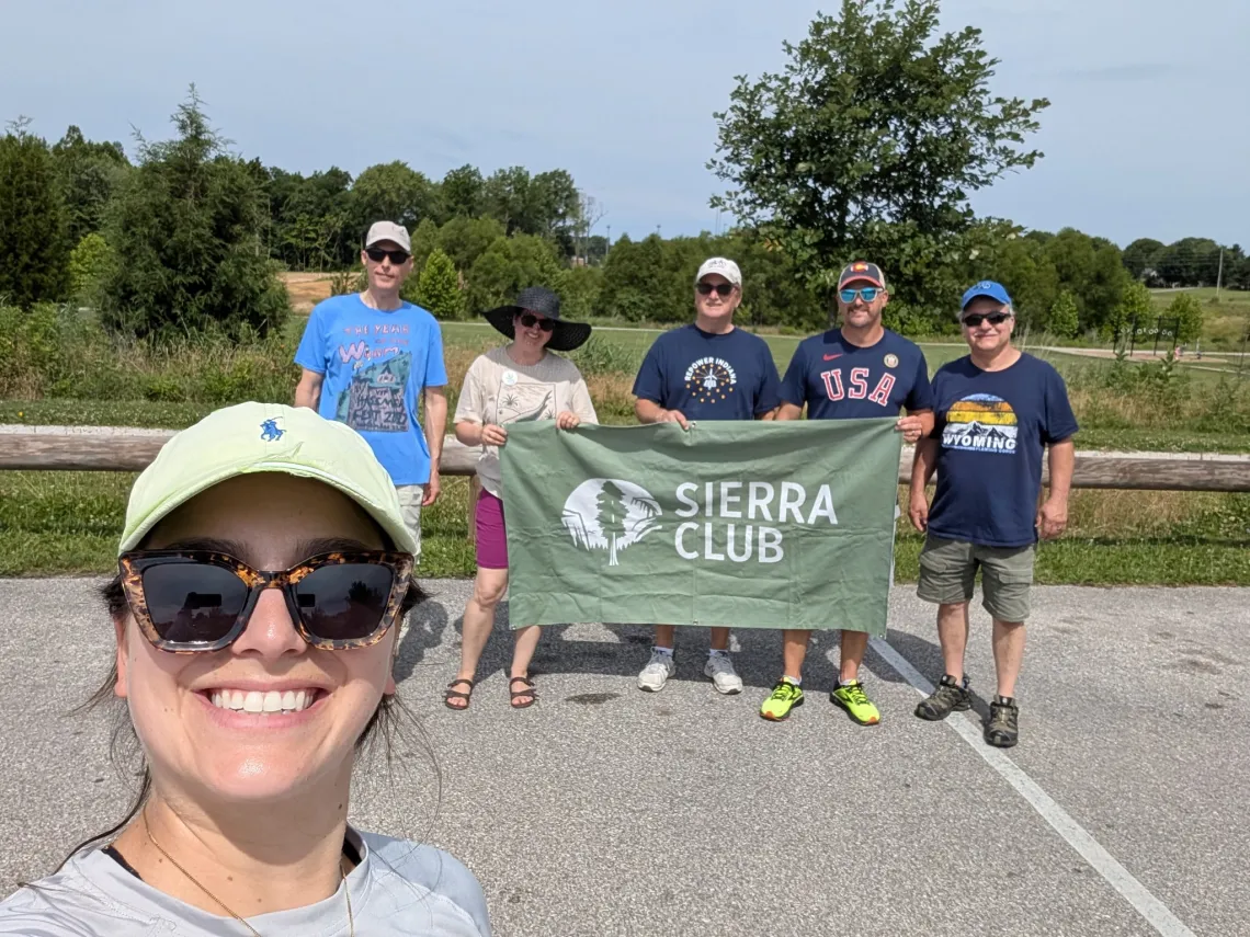 Five people in a parking lot with a park behind them with trails and trees and a hazy blue sky. The three folks in the middle are holding a green Sierra Club banner. In the foreground is the person taking the photo, selfie-style. She has a baseball cap and sunglasses and is smiling.