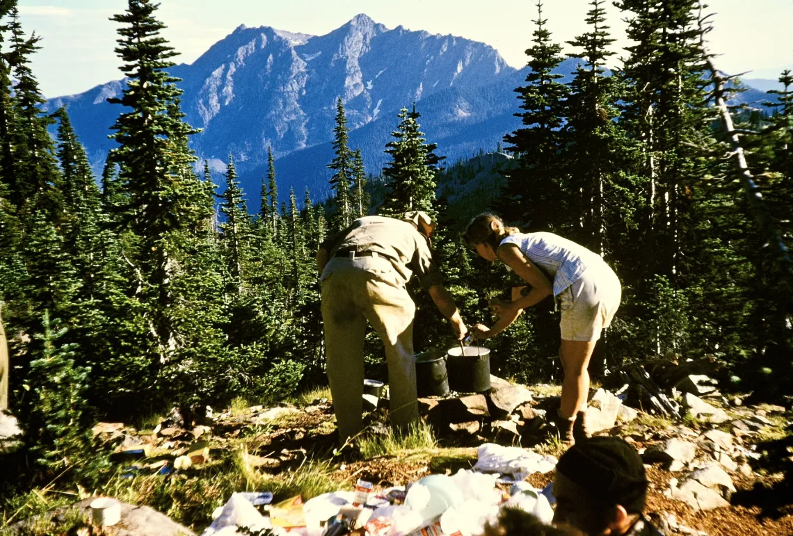a man and woman cook over a large camp pot on a rocky tarn surrounded by trees and mountains. Taken circa 1960s.