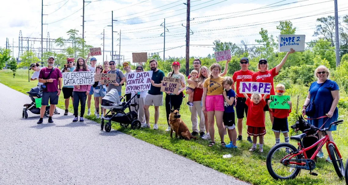 A group of around 20 people on a city trail. There is a paved trail path with grass to the side. They are holding signs to protest against a data center. A couple of them have bicycles. They are a mix of ages including some small children.