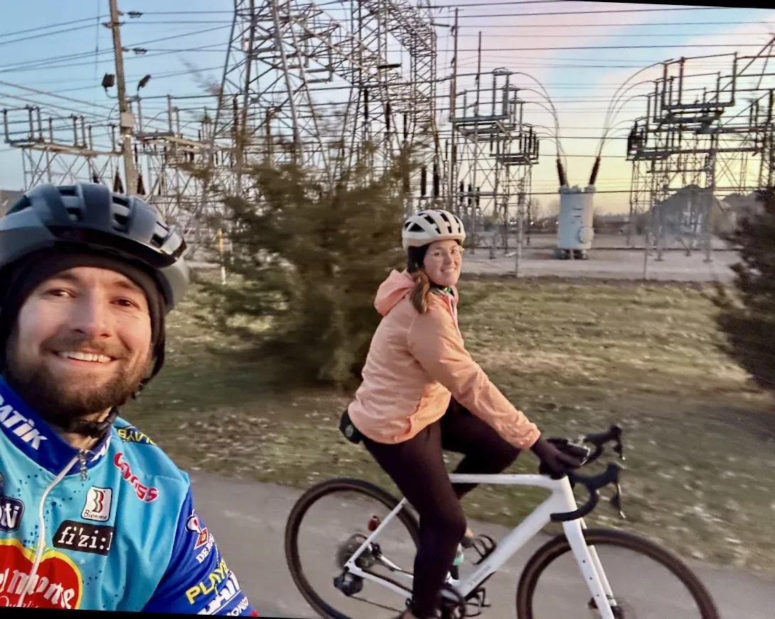 A selfie of someone on a bicycle with another person on a bike in shot too, both smiling and looking at the camera. They're riding on an urban trail and there is an electricity station in the background.