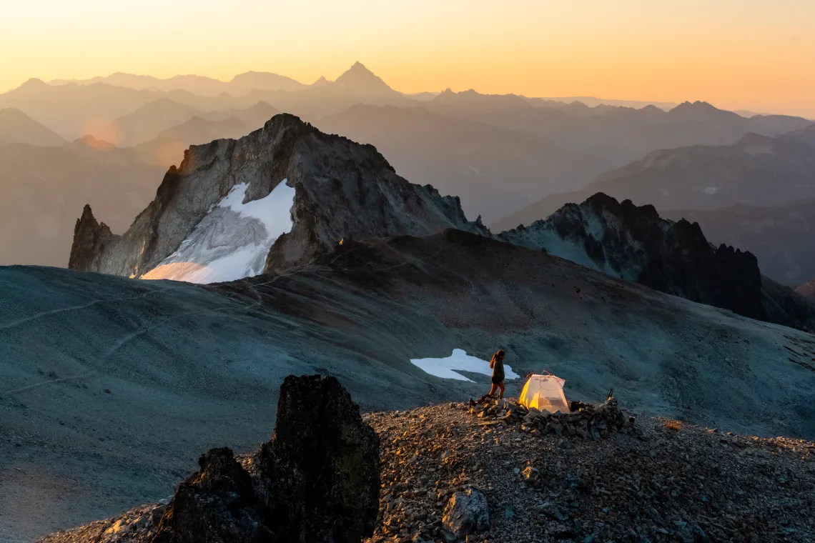 Layers of jagged mountain peaks, with a few small snowfields, under a soft yellow-orange sunset. There is a person with a tent perched on a small flat area in the foreground. 