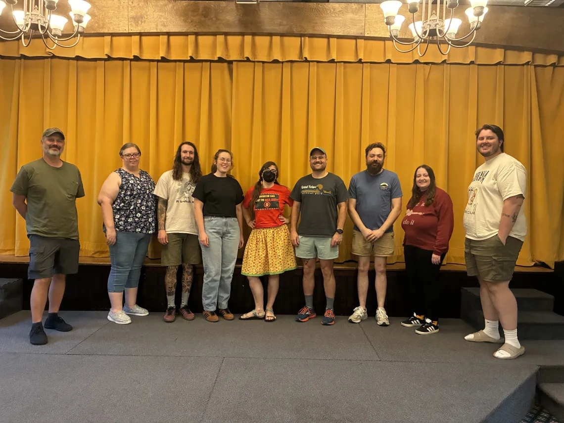 Nine people standing indoors in a church hall with a large mustard colored curtain behind them. They are all smiling for the photo. 