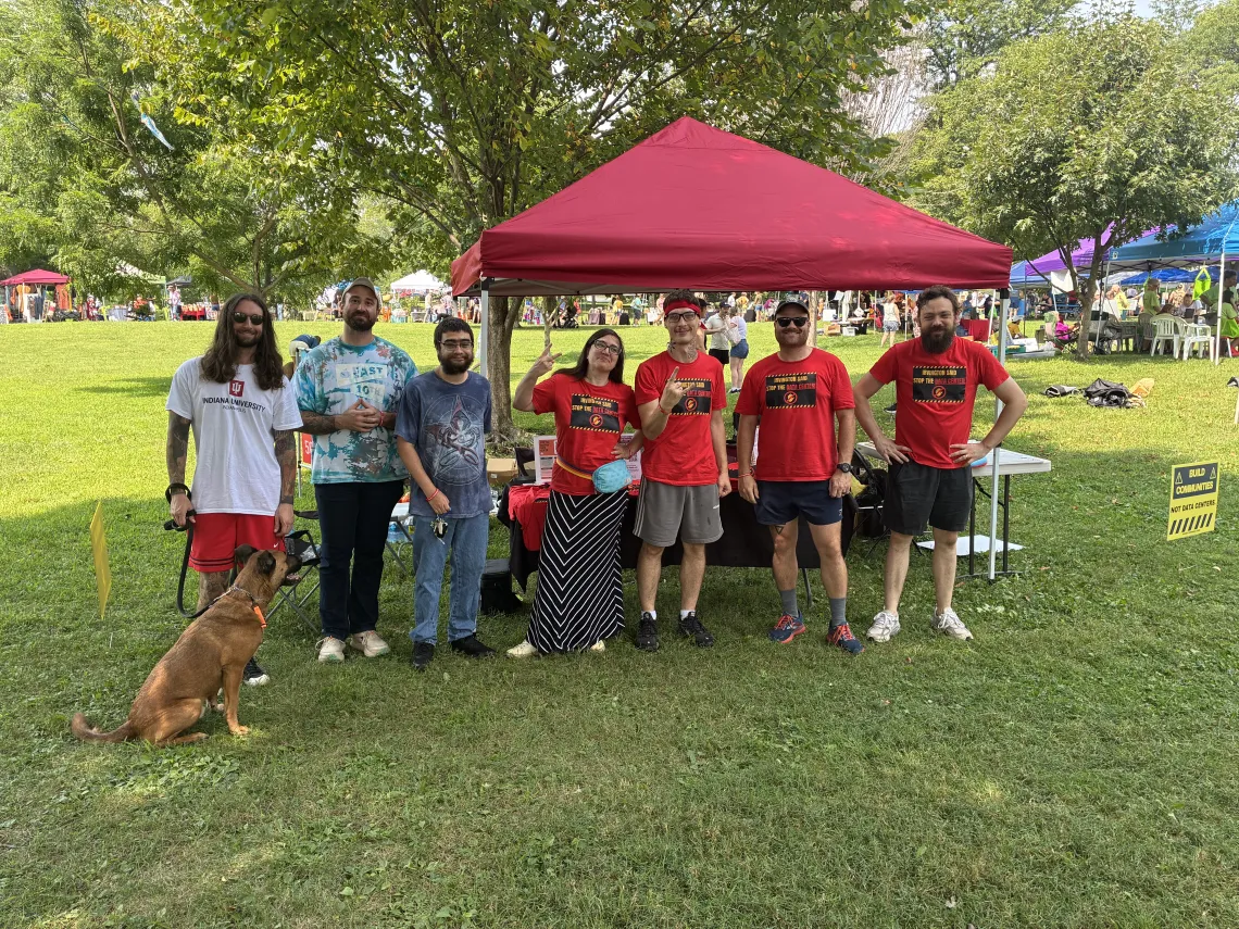 Seven people and a dog standing in front of a tabling tent in a park with trees and other tabling tents in the background, with folks milling around. Four of the people in the photo are wearing red t-shirts which say Irvington says Stop the Data Center