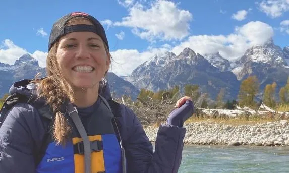 Stephanie Vermillion, a smiling young woman wearing a backward billed cap over her brown braid, stands in the left foreground near a rocky river bank with a jagged, mountainous horizon in the background under a white cloud studded blue sky.