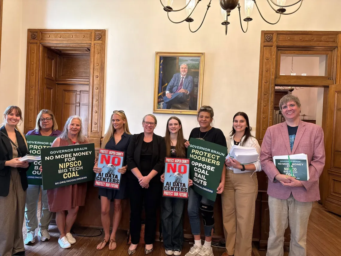 Nine people standing indoors, holding signs saying things like No AI data centers. Protect Hoosiers - oppose coal bail outs. Some of them are holding stacks of paper petitions.