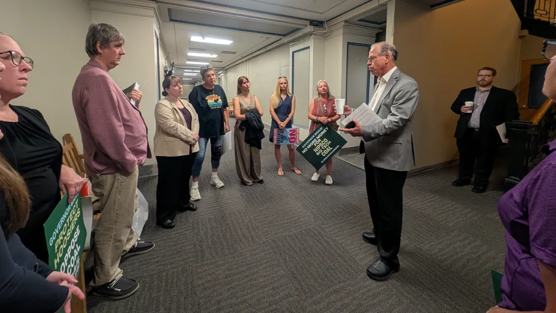 A group of around 11 people indoors in a lobby or corridor of an office. They are standing in a loose circle and talking to Gov Braun, a man who is wearing dark pants and a gray jacket and is holding some papers. Some of the folks have signs asking Gov. Braun to protect Hoosiers and oppose coal bail outs.
