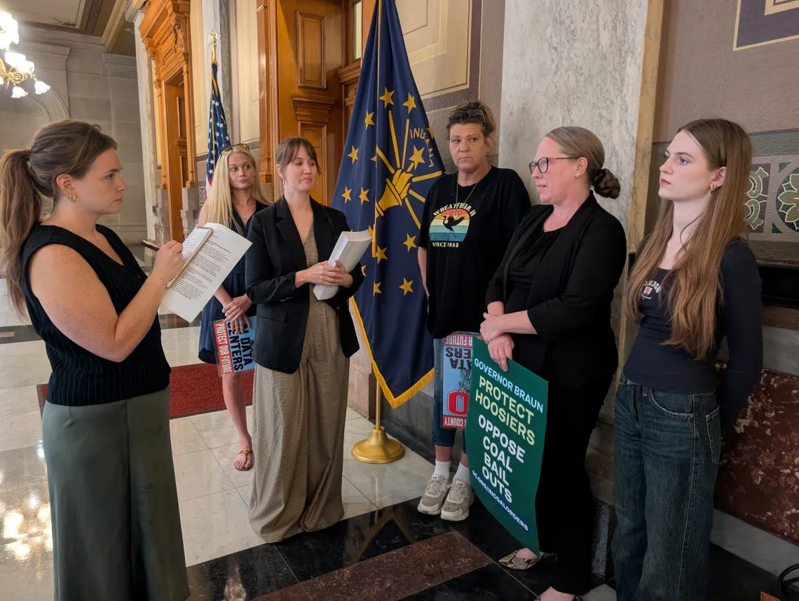 Six people standing inside the Statehouse building. There is an Indiana state flag. One of the people is interviewing two of the other people, one of which is holding a sign saying Governor Braun, Protect Hoosiers Oppose Coal Bail Outs