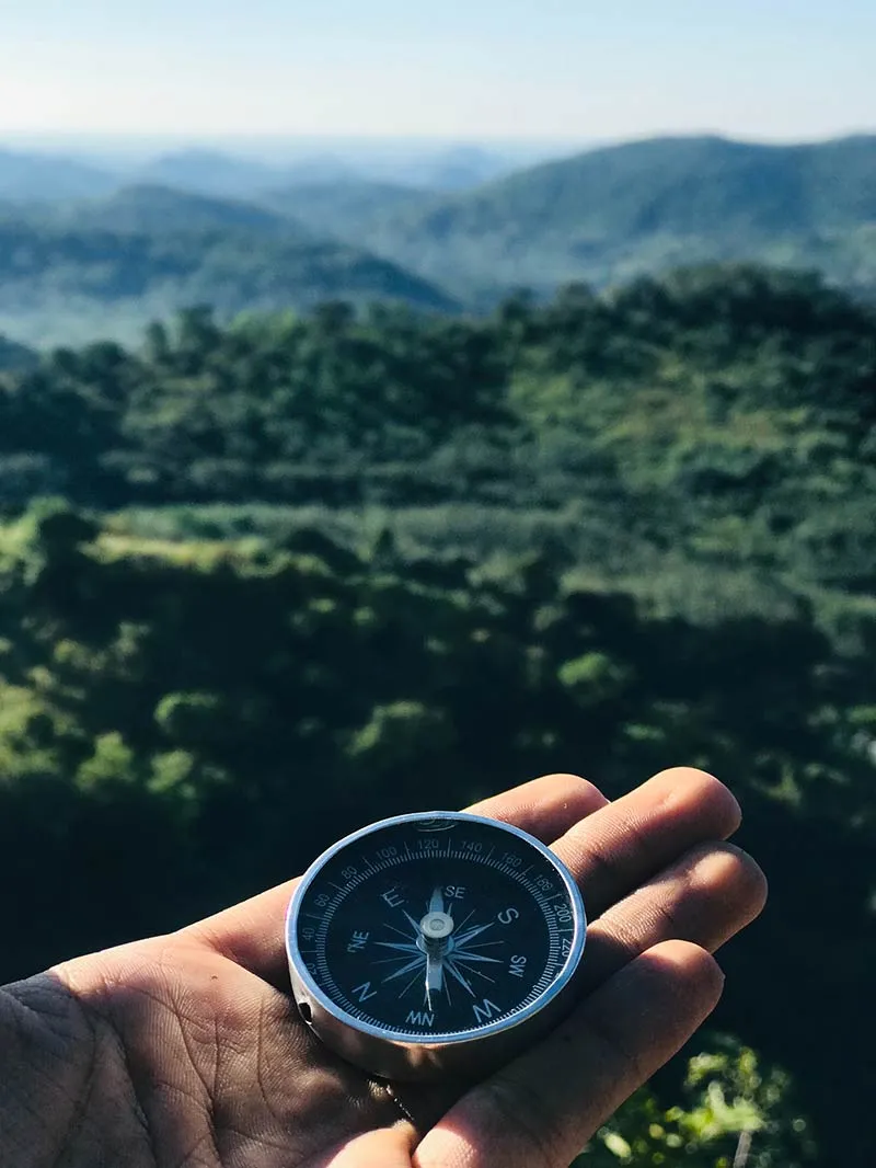 A hand holding a compass with hills and forest in the deep background.