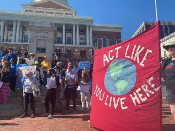 Rally in front of the MA State House with a large red banner that reads "Act like you live here" with a painting of the earth in the center.