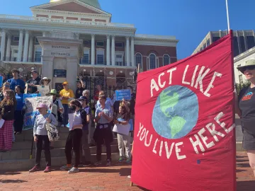 Rally in front of the MA State House with a large red banner that reads "Act like you live here" with a painting of the earth in the center.