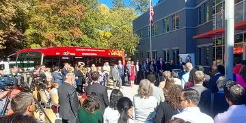 A wide-angle view of people watching Governor Cooper announce his executive order on clean transportation, outside ABB headquarters in Cary, NC. A red electric bus is in the background on this sunny day.