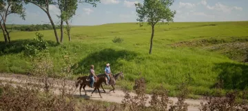 2 horseback riders on trail next to grassy field with trees