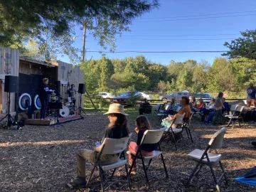 A young person stands on a small wooden stage at a microphone. There is a crowd of people seated on folding chairs and the ground is woodchip. The sky is blue and there are green trees in the background.